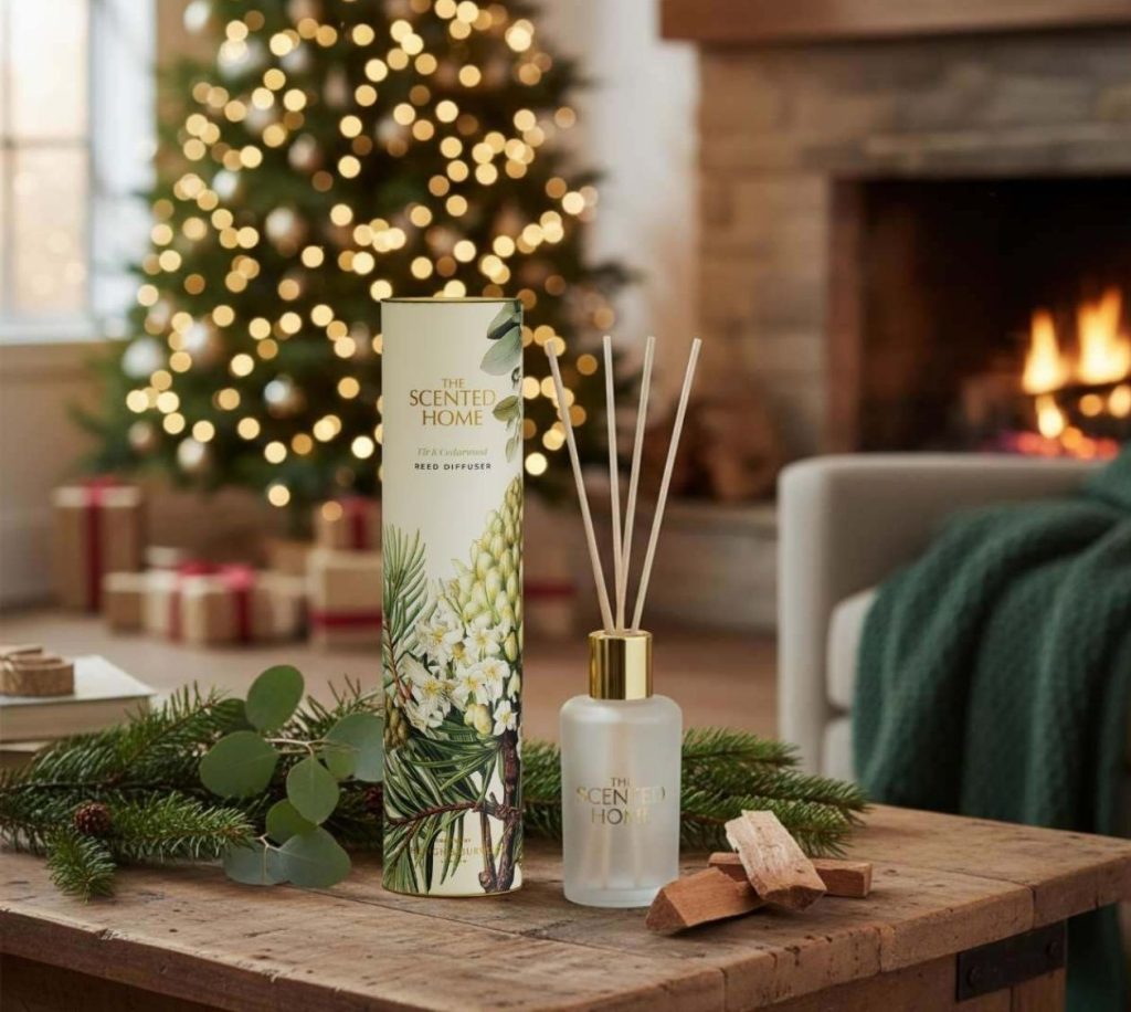 A Fir & Cedarwood reed diffuser by The Scented Home, surrounded by pine branches, wood chips, and a Christmas tree.