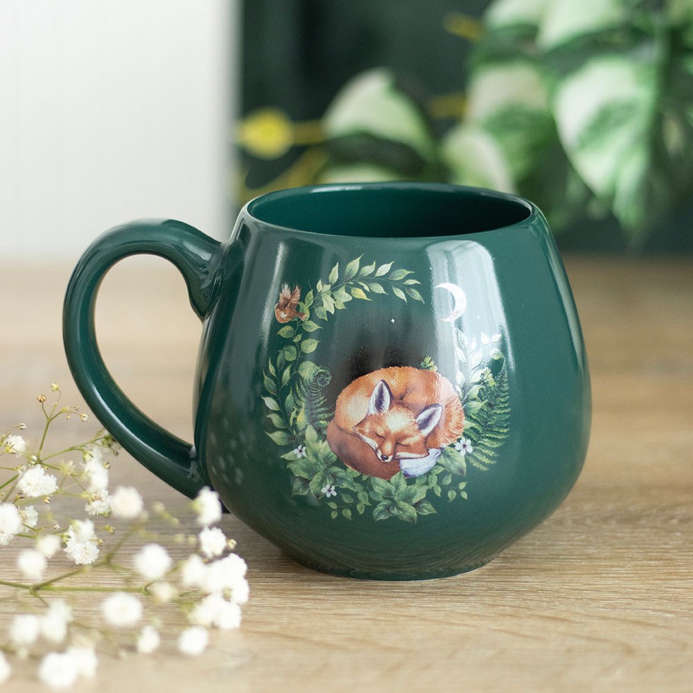 A dark green ceramic mug featuring a sleeping fox illustration surrounded by leaves, flowers, and a crescent moon.
