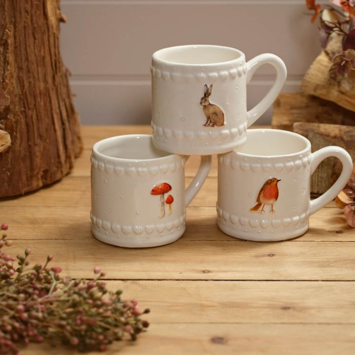 A set of three white ceramic mugs with embossed detailing, each featuring a countryside design – a rabbit, a red mushroom, and a robin.