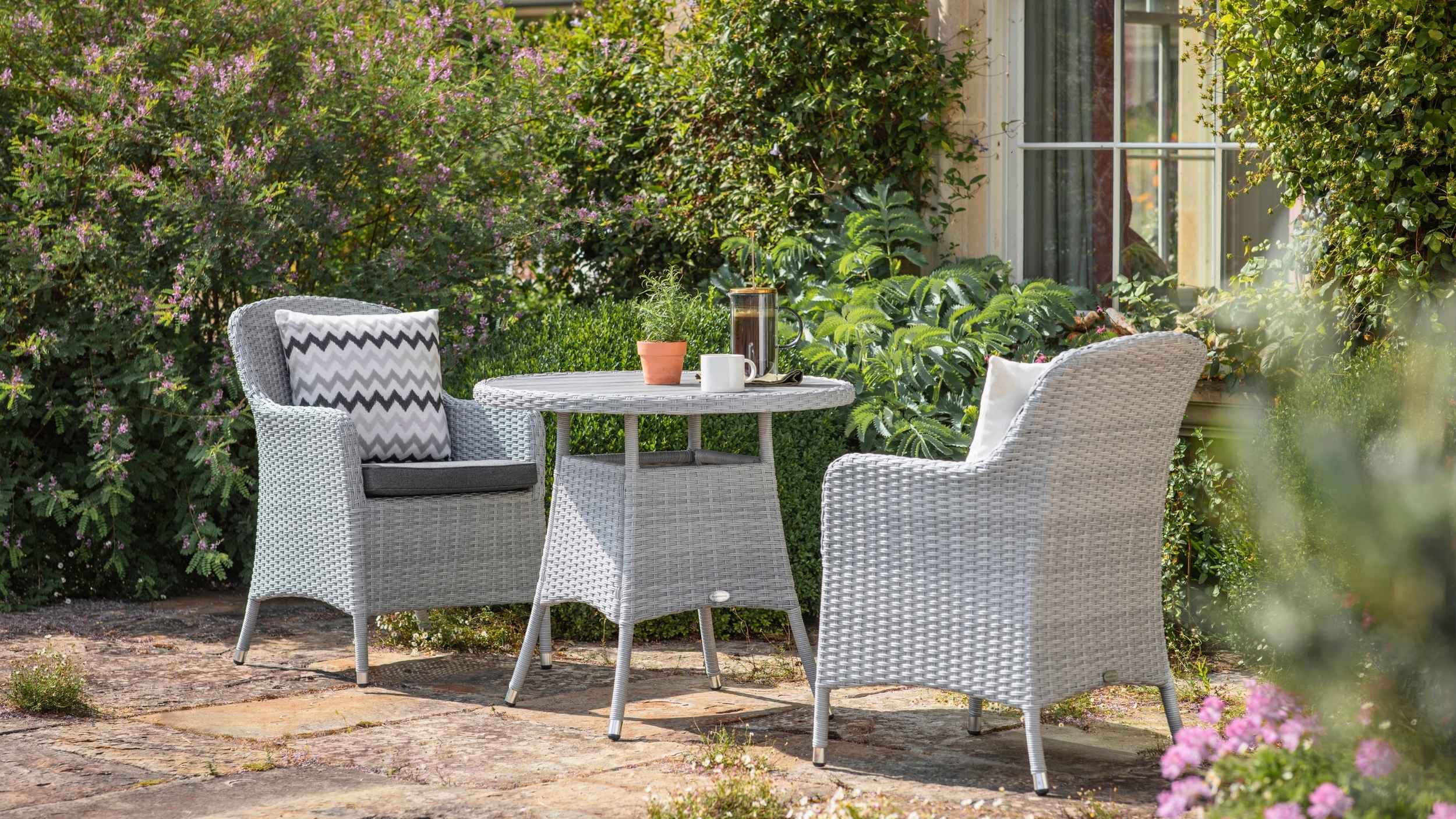 Bramblecrest Tetbury two seat bistro set in dove grey wicker with round table and cushioned armchairs on a sunlit garden patio.